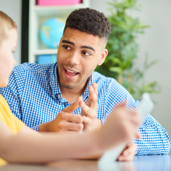 A teacher in a blue checkered shirt is engaged in conversation with a young student in a yellow shirt, gesturing with his hands as he speaks attentively.