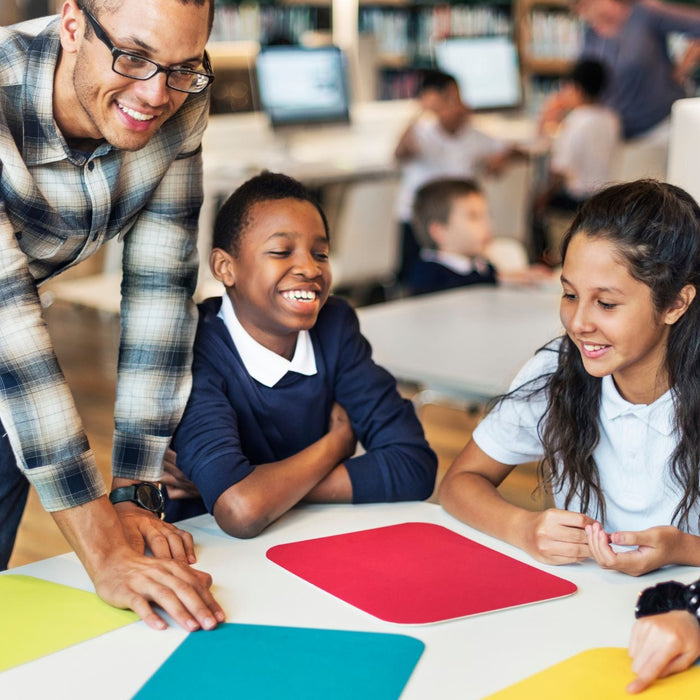 A teacher leans over a table, smiling as he engages with a group of students who are also smiling and interacting with colorful paper squares in a classroom setting.