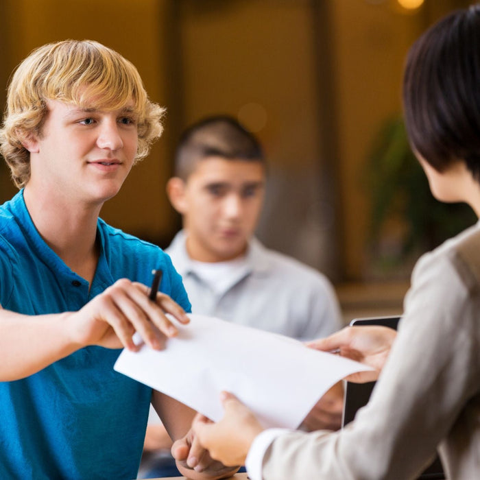 A young man in a blue shirt hands a document to a woman seated across from him, while other students are seen working in the background.