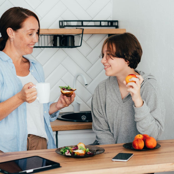 A woman and a teenager share a moment in a modern kitchen. The woman holds a coffee mug and a piece of toast with greens, while the teenager holds a peach, smiling at each other. A plate of peaches, a salad, and a tablet are on the counter in front of the