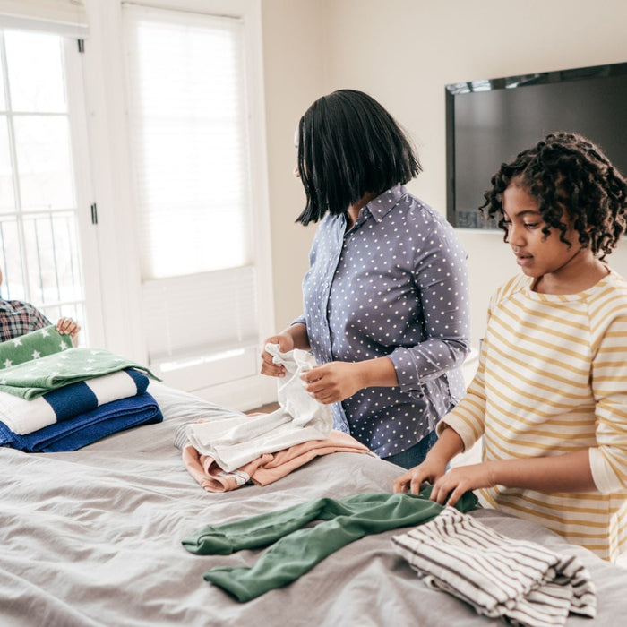 A mother and her two children are folding laundry together in a bright bedroom with large windows, as neatly folded clothes are stacked on the bed. The younger boy smiles while holding a folded green towel, while the older girl carefully folds a striped s