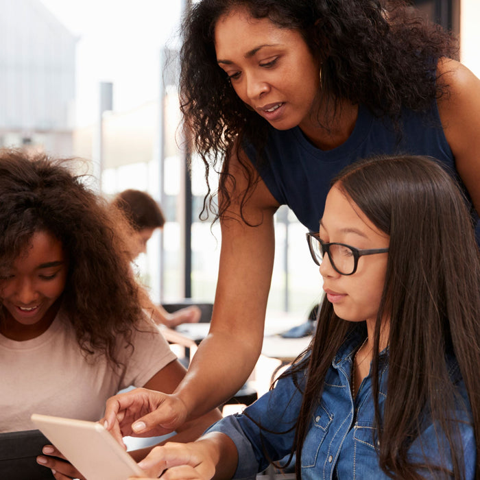 A teacher leans over to assist two students with their tablets, guiding them as they focus on their screens in a classroom setting.