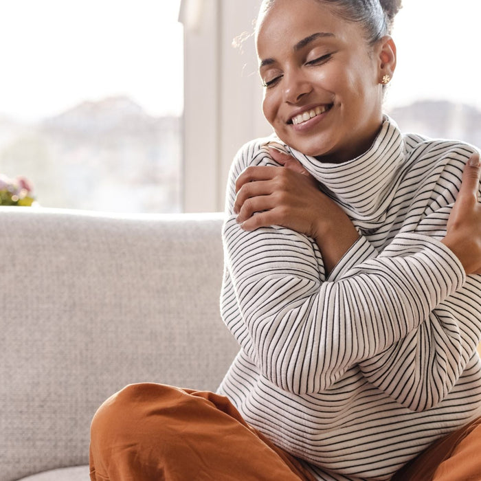 A woman sits cross-legged on a sofa, smiling with her eyes closed as she hugs herself, conveying a sense of warmth and self-love.